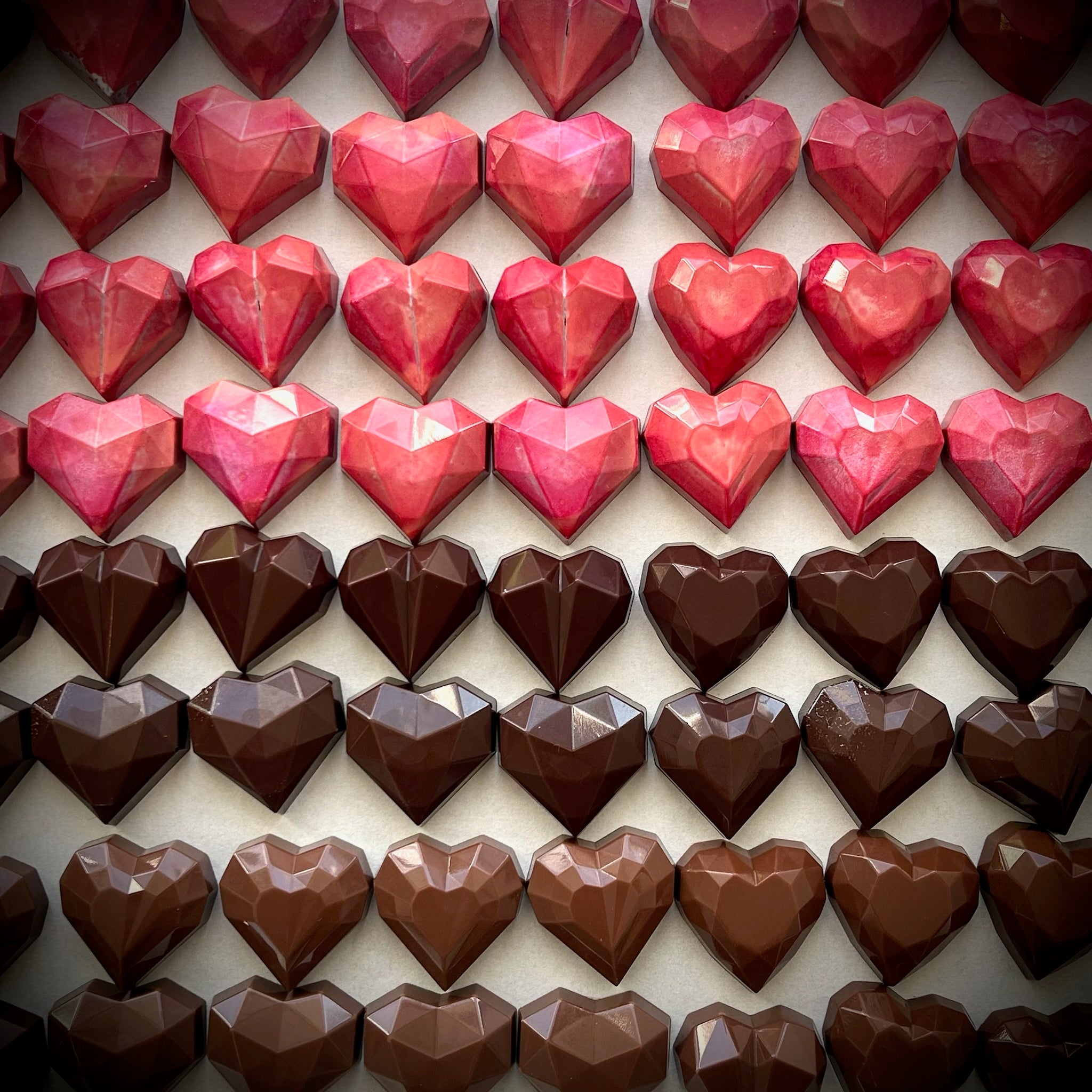Chocolats artisanaux rouges et bruns en forme de cœur, alignés en rangées. Chaque coffret contient 2 cœurs caramel, 2 cœurs praliné maison, et 2 cœurs gelée d’airelle et praliné amandes. Fabriqué à Montréal, idéal pour la Saint-Valentin.
