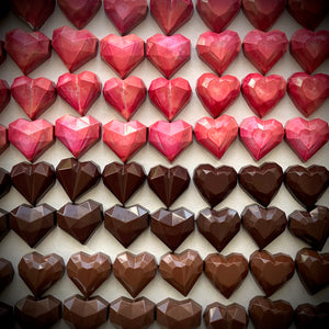 Chocolats artisanaux rouges et bruns en forme de cœur, alignés en rangées. Chaque coffret contient 2 cœurs caramel, 2 cœurs praliné maison, et 2 cœurs gelée d’airelle et praliné amandes. Fabriqué à Montréal, idéal pour la Saint-Valentin.